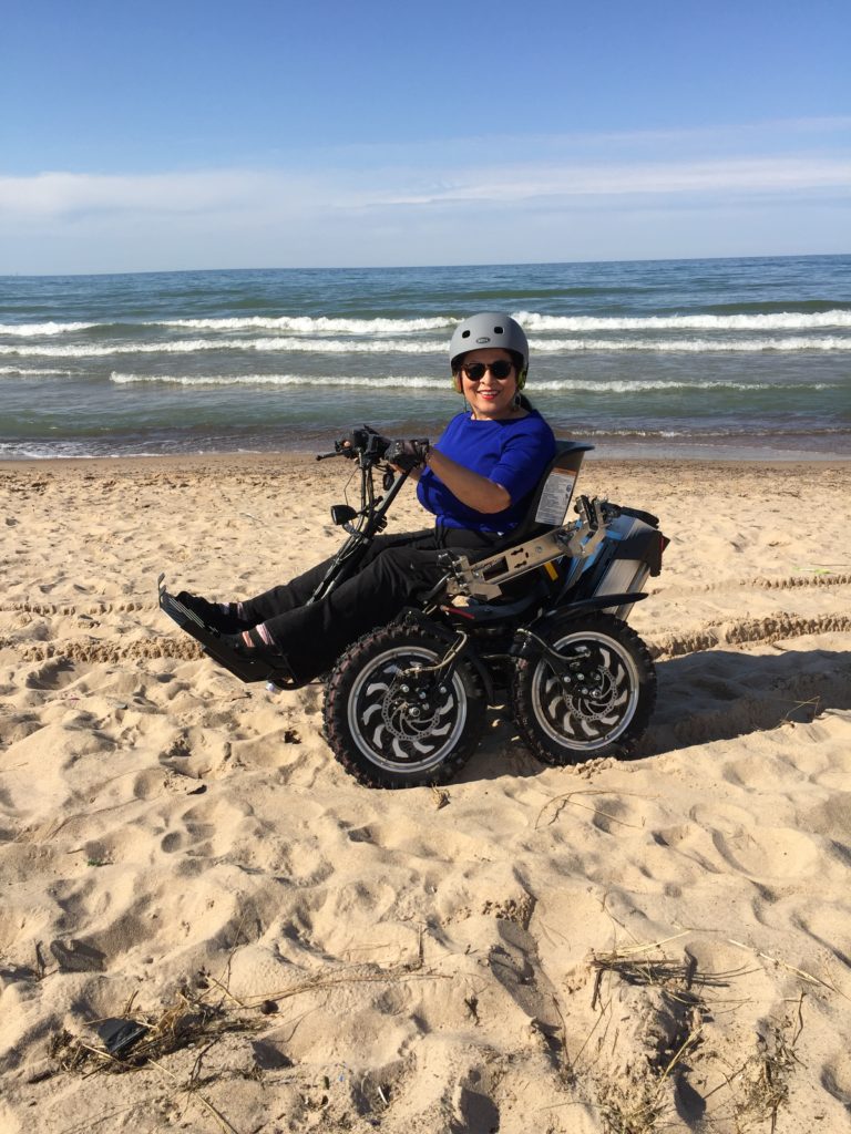 demonstration of the Zoom chair on the sandy beach by Zully wearing a helmet on head with sunglasses hands on controls and legs extended on foot pedals 