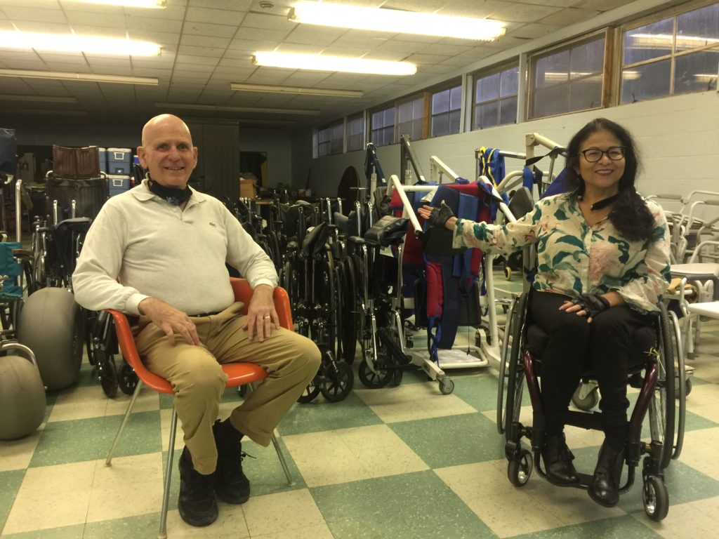 Bob sitting in orange chair and Zully sits in her wheelchair pointing to room filled with durable medical equipment in back of them.